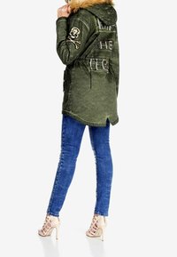 Parka vert olive avec une capuche en fausse fourrure, un patch crâne et os croisés, et des accents de fermeture éclair ; associé à un jean bleu ajusté et des sandales à talons beiges.