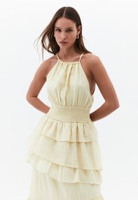 Žluté šatovky s tiered designem, smocked pasem, halter krkem a vrstvenými volány. Vyrobeno z lehké tkaniny s měkkou strukturou.