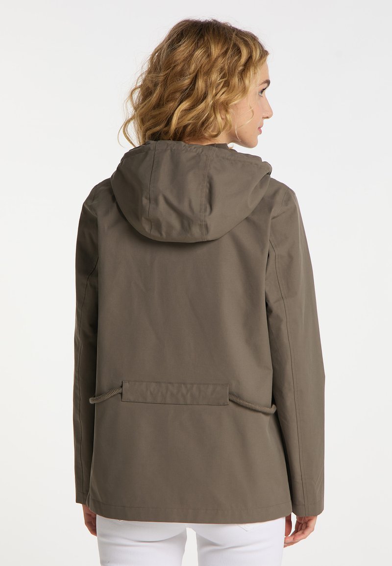 Vrouw met krullend blond haar, gekleed in een bruine hoodie en witte broeken, van achteren zichtbaar tegen een neutrale achtergrond.