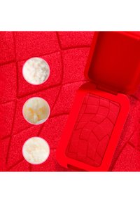 Trois petits contenants avec de la crème à côté d’un étui compact rouge ouvert, posés sur une surface rouge texturée avec des motifs géométriques.