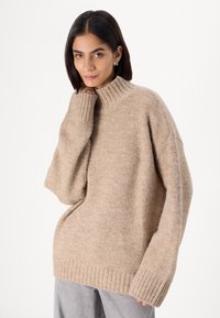Beige knit sweater with a high ribbed neckline, oversized fit, and wide sleeves. Textured fabric with a soft appearance.