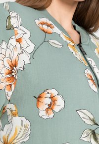 Chaqueta bomber floral en un tejido verde apagado. Presenta grandes flores naranjas y blancas, cierre frontal de cremallera y cuello acanalado. Textura suave.