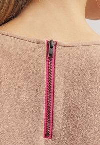 Detailní záběr na zip v zadním výstřihu na béžové texturované látce s viditelnou růžovou zipovou páskou.