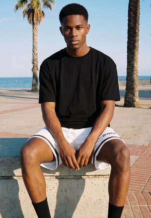 Jeune homme en t-shirt noir et short blanc assis sur un banc en béton près des palmiers au bord de la mer sous un ciel clair.