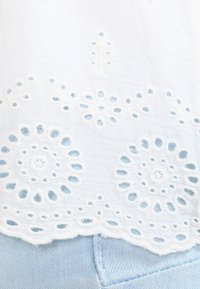 Tissu en coton blanc avec des motifs floraux brodés et des détails en œillet. Le design présente un bord festonné, sur fond de denim bleu clair.