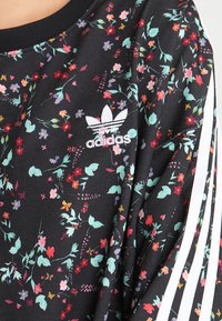 Tela negra con un patrón floral colorido, con un logo de Adidas bordado en blanco y dos franjas blancas a lo largo de la manga.