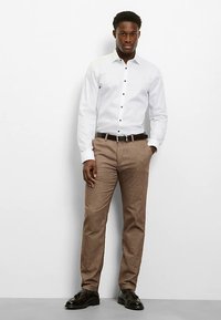 Weißes Langarmhemd mit schwarzen Knöpfen, kombiniert mit brauner Strukturhose und einem schwarzen Gürtel. Das Modell steht selbstbewusst vor einem neutralen Hintergrund.