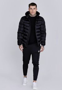 Sort dunjakke med vandret quiltning, lynlås og hætte. Båret over en sort sweater og kombineret med sorte tilspidsede joggingbukser og hvide sneakers.