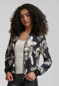 Veste bomber noire avec motif de feuilles de palmier blanches et brunes, fermeture éclair, poignets côtelés et col, portée par-dessus un débardeur clair.