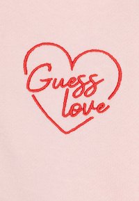 Lys rosa stoff prydet med et rødt brodert design formet som et hjerte, som viser teksten "Guess love" i en kursiv skrifttype.