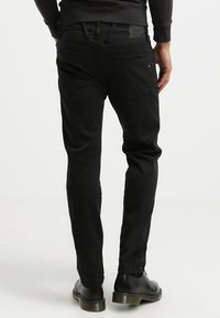 Svart slim-fit jeans med en slät textur, med bakfickor och en etikett vid midjan, kombinerat med svarta skor.