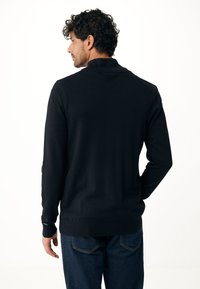 Svart långärmad tröja med hög krage, tillverkad av mjukt tyg. Har ribbade ärmslut och nederkant, visas bakifrån med mörka denimjeans.