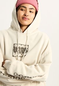 Crème-kleurige hoodie van zachte stof, met een grafische print op de voorkant en een intricaat ontwerp langs de mouwen. Inclusief een capuchon.