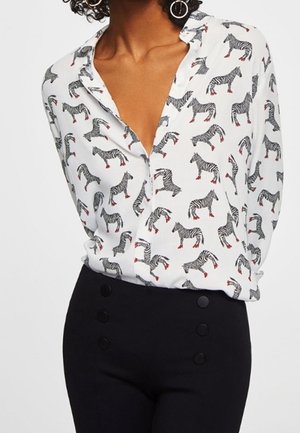 Femme portant une chemise blanche à boutons avec des imprimés zèbre noirs et des sabots rouges, associée à un pantalon noir taille haute avec six boutons.