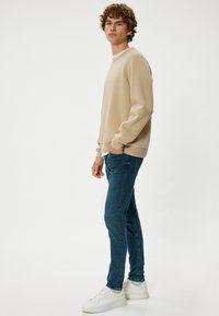 Vaaleanbeige collegepaita, jossa on pitkät hihat ja resorireunat, yhdistettynä slim-fit tummansinihin farkkuihin ja valkoisiin tennareihin. Yksinkertainen muotoilu, ei näkyviä kuvioita.