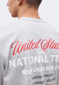 Άντρας που φορά γκρι πουκάμισο με κόκκινο και λευκό κείμενο που γράφει "United States Men's National Team" και επιπλέον μικρότερο τυπωμένο κείμενο.