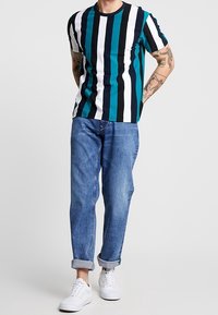 Camiseta de manga corta a rayas en tonos verde azulado, negro y blanco, combinada con vaqueros azul enrollados en el dobladillo y zapatillas blancas. Corte casual y mezcla de algodón.