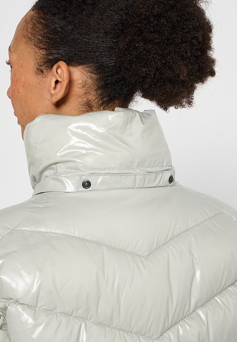 Hellgraue, glänzende Steppjacke mit hohem Kragen, ausgestattet mit Druckknöpfen und gestepptem Zickzackmuster auf der Rückseite.