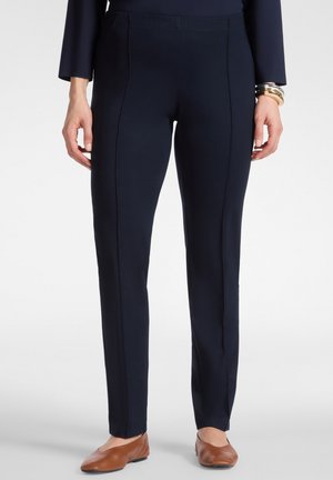 Vrouw draagt zwarte slim-fit broek en bruine platte schoenen, met armbanden om haar rechterpols, staand tegen een effen achtergrond.