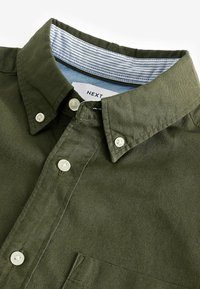 Camicia verde oliva con bottoni bianchi, un colletto interno a righe e una piccola tasca sul petto.