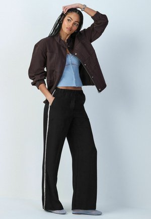 Woman with long braided hair wearing a brown jacket, light blue crop top, black pants with white trim, and blue mesh shoes, posing with hand on head.