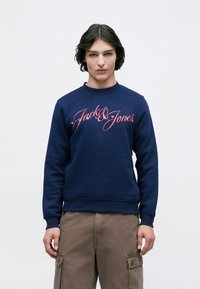 Mladý človek s tmavými stredne dlhými vlasmi, oblečený v námorníckej mikine s červeným logom "Jack & Jones" a hnedých cargo nohaviciach, stojí na bielom pozadí.