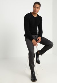 Black ribbed long-sleeve top paired with dark gray slim-fit trousers. Smooth texture, ankle-height black boots. Seated on a white chair.