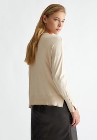 Maglione beige lavorato a maglia con maniche lunghe, caratterizzato da un orlo grezzo e tre bottoni decorativi su ogni manica. Materiale morbido e vestibilità rilassata.