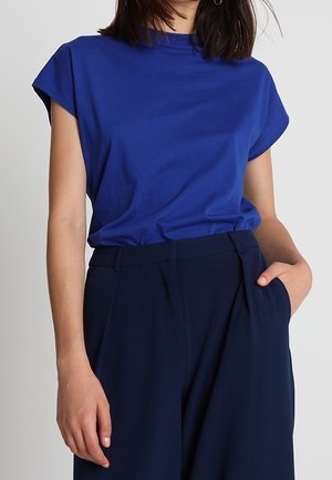 Sininen lyhythihainen puuvillapusero, jossa on korkea kaulus, yhdistetty tummansinisiin korkeavyötäröisiin housuihin. Kangas näyttää sileältä ja mukavalta.