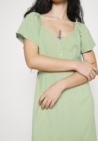 Vaaleanvihreä mekko, jossa on lyhyet puffihihat, V-mallinen kaula-aukko ja napitus edessä. Pehmeä kangas rakenne, jossa on hillitty kiilto.