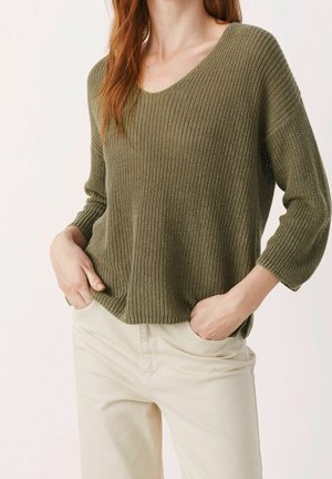 Nainen, jolla on päällään oliivinvihreä ribbineulottu V-kaula-aukollinen neulepusero, jossa on 3/4-hihat, ja vaaleanbeiget housut, kädet rennosti taskuissa.