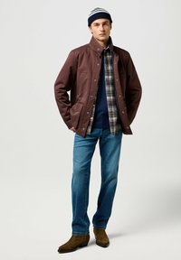 Chaqueta de nylon marrón, abotonada, con una camisa de cuadro debajo. Jeans azul claros y botas marrones. Gorro con rayas azules y blancas.