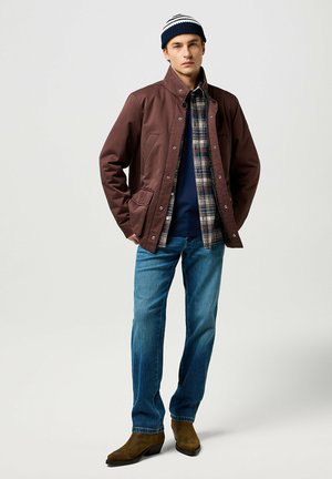 Chaqueta de nylon marrón, abotonada, con una camisa de cuadro debajo. Jeans azul claros y botas marrones. Gorro con rayas azules y blancas.