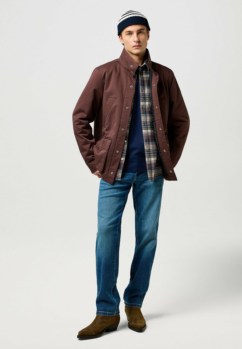 Chaqueta de nylon marrón, abotonada, con una camisa de cuadro debajo. Jeans azul claros y botas marrones. Gorro con rayas azules y blancas.
