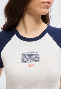 Femeie purtând un tricou raglan alb cu albastru închis, cu radio vintage imprimat și logo roșu Levi's pe piept, privind pe lateral.