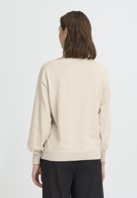 Bézs pulóver laza szabással, bordázott mandzsettákkal és széles nyakkivágással. Puha textúra és minimális varrásdíszítés. Fekete nadrággal stylingolva.