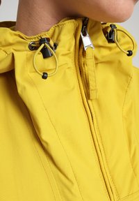 Geel jack met hoge kraag en trekkoorden, ritsluiting en zichtbare stiknaden langs de naden. Gladde, duurzame stoftextuur.