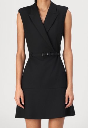 Vestido negro sin mangas con cuello en V, hombreras y un cinturón negro fino en la cintura, llevado por una persona de pie.