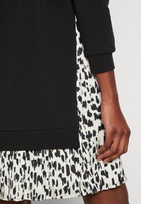 Schwarzes Baumwolloberteil mit langen Ärmeln, kombiniert mit einem weißen plissierten Rock mit einem schwarz-weißen abstrakten Muster. Sichtbare Nähte und Branding-Details.