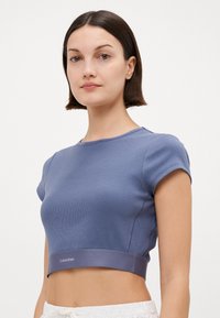 Zkrácený žebrovaný tričko v tlumené modré barvě s krátkými rukávy a hladkým elastickým páskem na lemu, na němž je logo Calvin Klein.