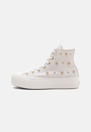 Fehér magas szárú tornacipő vastag talppal, arany szemekkel és díszítő arany szegecsekkel vászon felsőrészen, oldalnézet halvány háttéren.