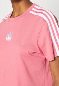 Rosa bomullströja med korta ärmar, rund halsringning, vita randiga detaljer och en blå och vit logotyp på bröstet.