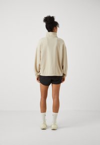 Pullover beige con colletto alto e vestibilità rilassata, abbinato a pantaloni corti neri. Il modello indossa calze bianche e sneakers color crema.