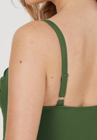 Mörkgrön texturerad tyg med tunna justerbara band, som visar en detaljerad ryggdesign. Inkluderar metallkomponenter för justering av banden.