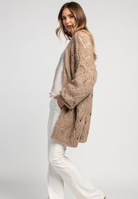 Beżowy sweter kardigan o luźnym kroju, z teksturowanym otwartym wzorem. Nosi się go na jasnoszary top i białe spodnie, w stylu casualowym.