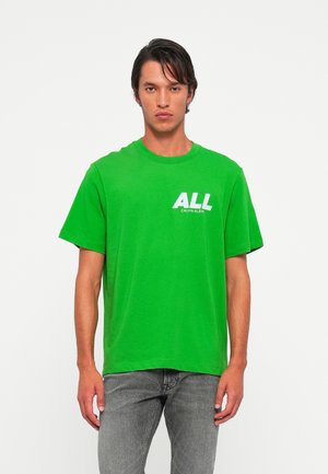 Man wearing bright green Calvin Klein T-shirt with "ALL" logo, paired with gray jeans, standing against plain white background.