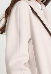 Světlý béžový kabát z vlněné směsi s klopovým límcem, volným střihem a jemnou strukturou. Má široké rukávy a minimální detaily švů.