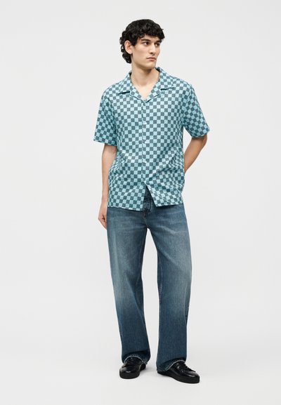 Νεαρός άνδρας που φοράει μπλε και μαύρο καρό κοντομάνικο πουκάμισο, μπλε τζιν και μαύρα παπούτσια, στέκεται με το ένα χέρι πίσω από την πλάτη του.
