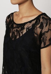 Svart spetsblus med blommönster; avslappnad passform; har korta ärmar och en vid halsringning; semi-genomskinlig textur som kontrasterar med en ogenomskinlig bas.