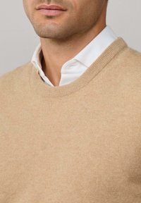 Svetlo bež lagani pleteni pulover s okruglim izrezom, koji ima mekanu teksturu i tanke rebraste naglaske na ovratniku. Ispod se vidi bijela košulja.